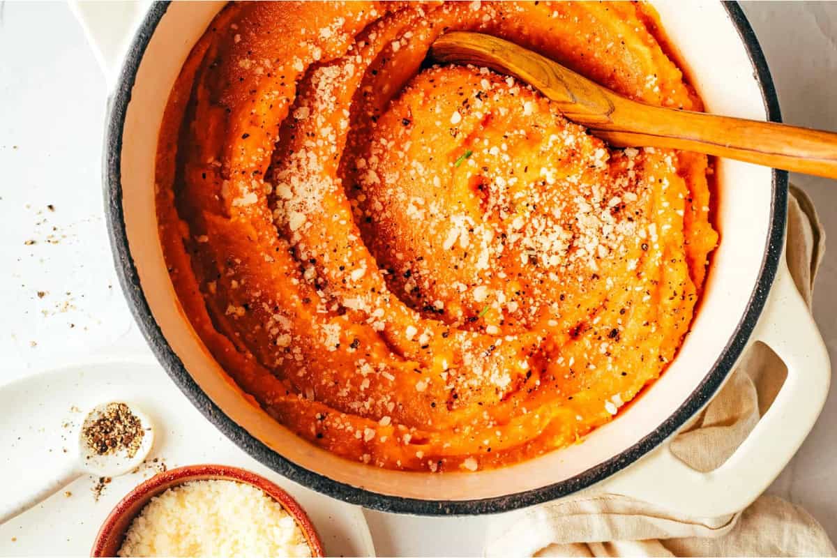 A pot of creamy orange mashed sweet potatoes topped with grated parmesan cheese and black pepper. A wooden spoon is immersed in the mash, and a small dish of additional cheese and pepper is placed nearby.