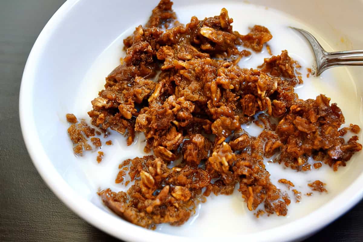 A bowl of granola cereal with milk, featuring clusters of oats and nuts. A spoon is partially submerged in the milk and granola mixture. The bowl is placed on a dark surface.