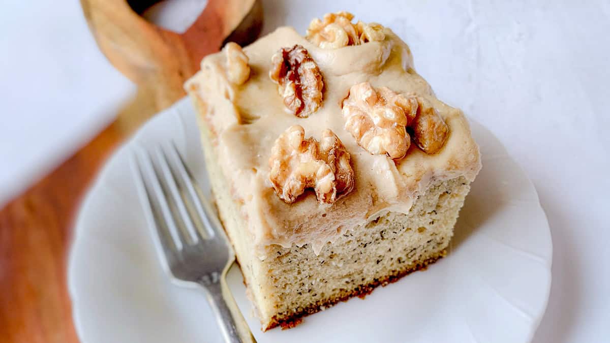 A slice of banana cake topped with thick frosting and walnut halves on a white plate. A fork is placed beside the cake. The background features a wooden ring and a white surface.
