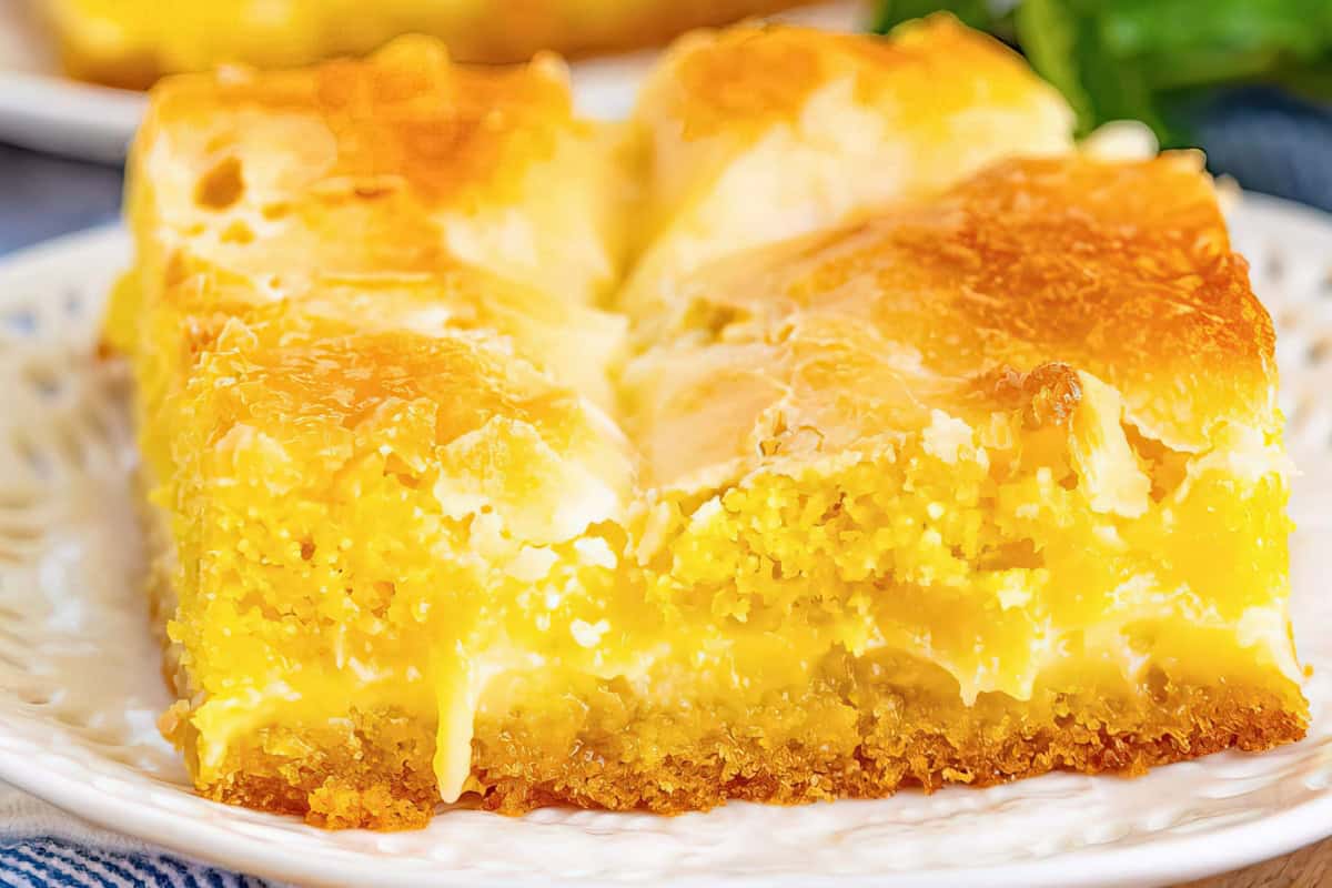 A close-up of a slice of gooey butter cake with a golden-brown top and a moist, dense yellow interior. The dessert is served on a white plate, showcasing its creamy and crumbly texture.