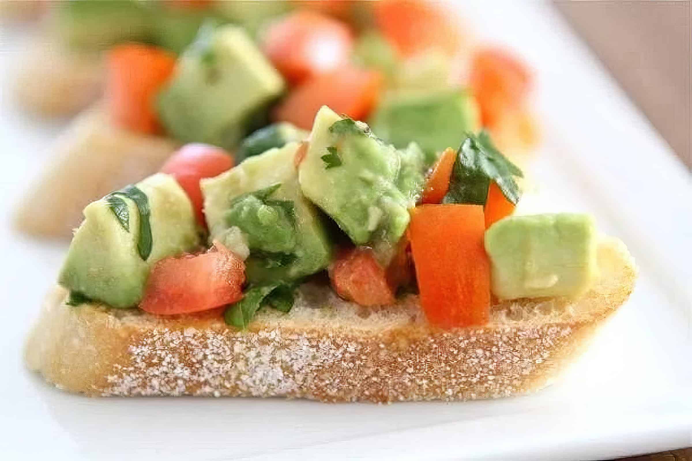 A piece of crusty bread topped with chopped avocado, tomato, and fresh herbs sits on a white plate. The colors are vibrant, and the texture of the ingredients contrasts with the crispness of the bread.