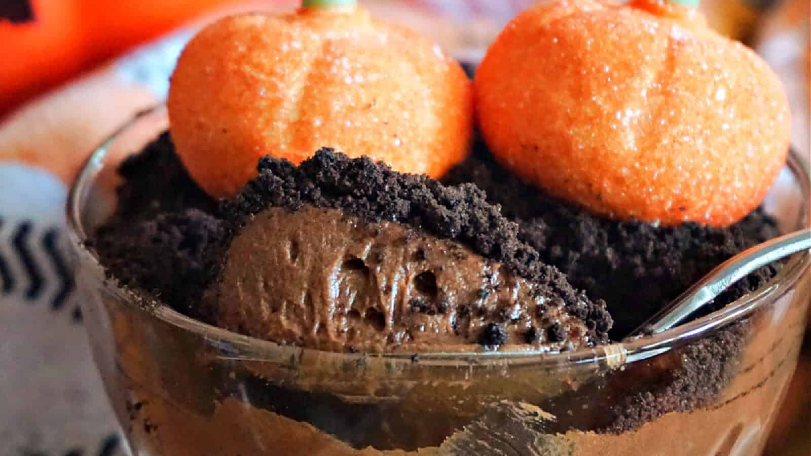 A dessert cup filled with chocolate pudding is topped with crushed cookies and two orange pumpkin-shaped candies. A spoon is partially visible, digging into the pudding. Cozy, blurred background enhances the autumn or Halloween theme.