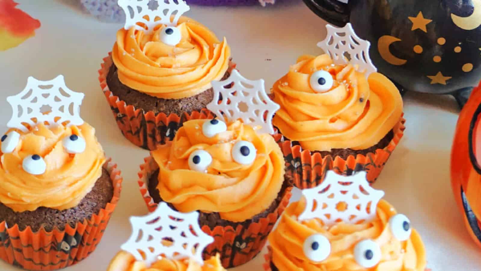 Halloween-themed cupcakes with orange frosting, topped with edible eyes and white spiderweb decorations. The cupcakes are in orange and black wrappers, surrounded by festive Halloween decor.