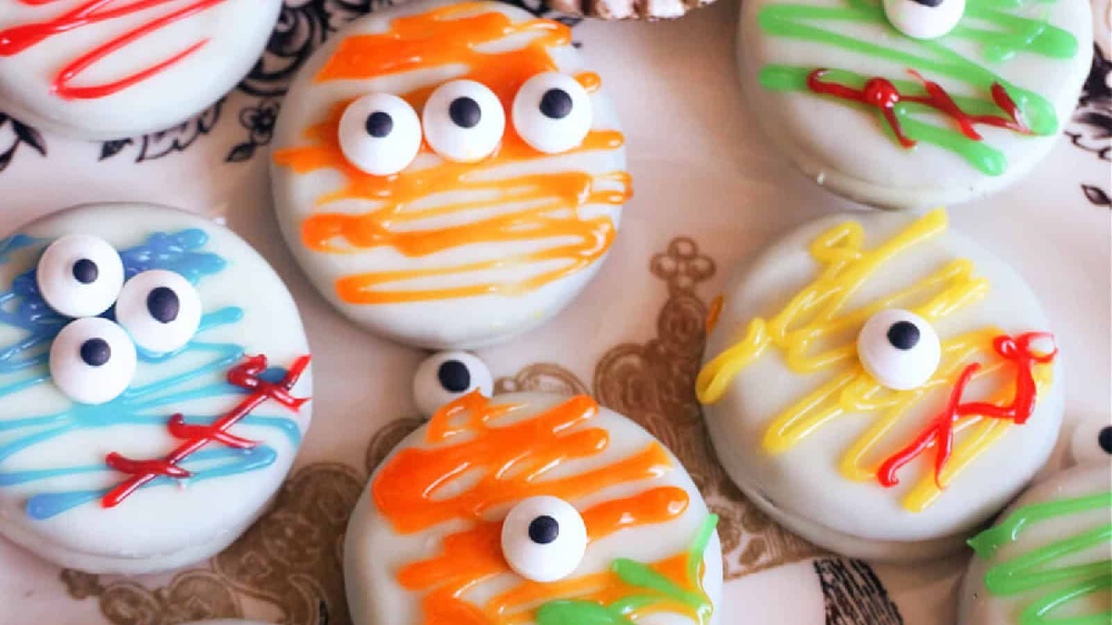 Colorful Halloween-themed cookies with white icing topped with candy eyes. Each cookie is decorated with striped patterns in orange, yellow, blue, and green, resembling playful monsters or mummies.