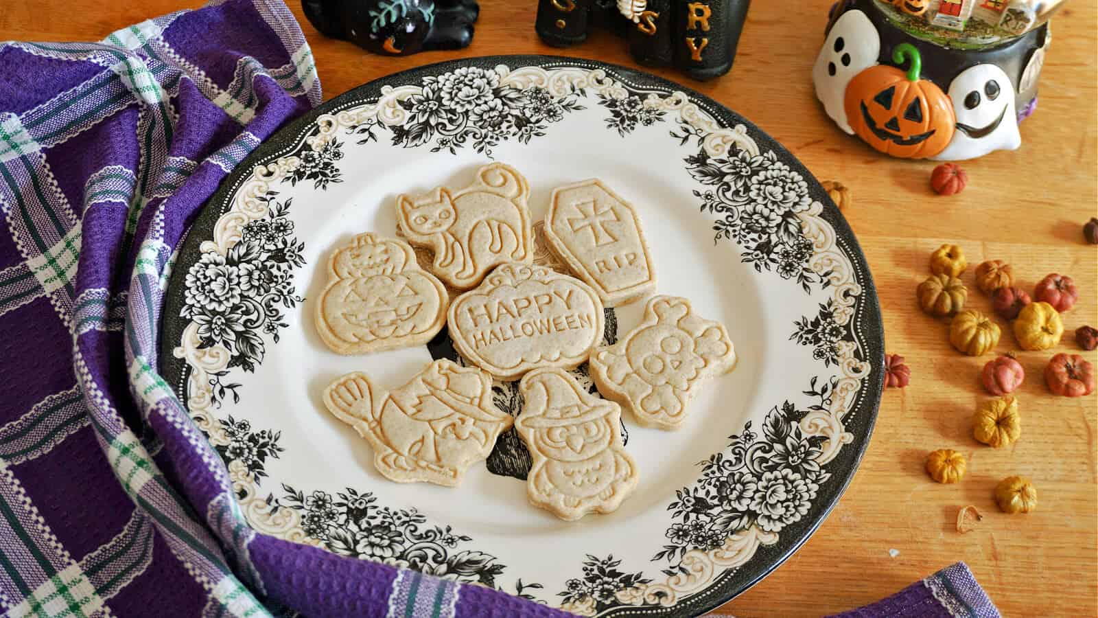 A decorative plate displays Halloween-themed cookies shaped like a cat, tombstone, pumpkin, and witch. It's surrounded by a purple plaid cloth and small autumn decorations on a wooden table.
