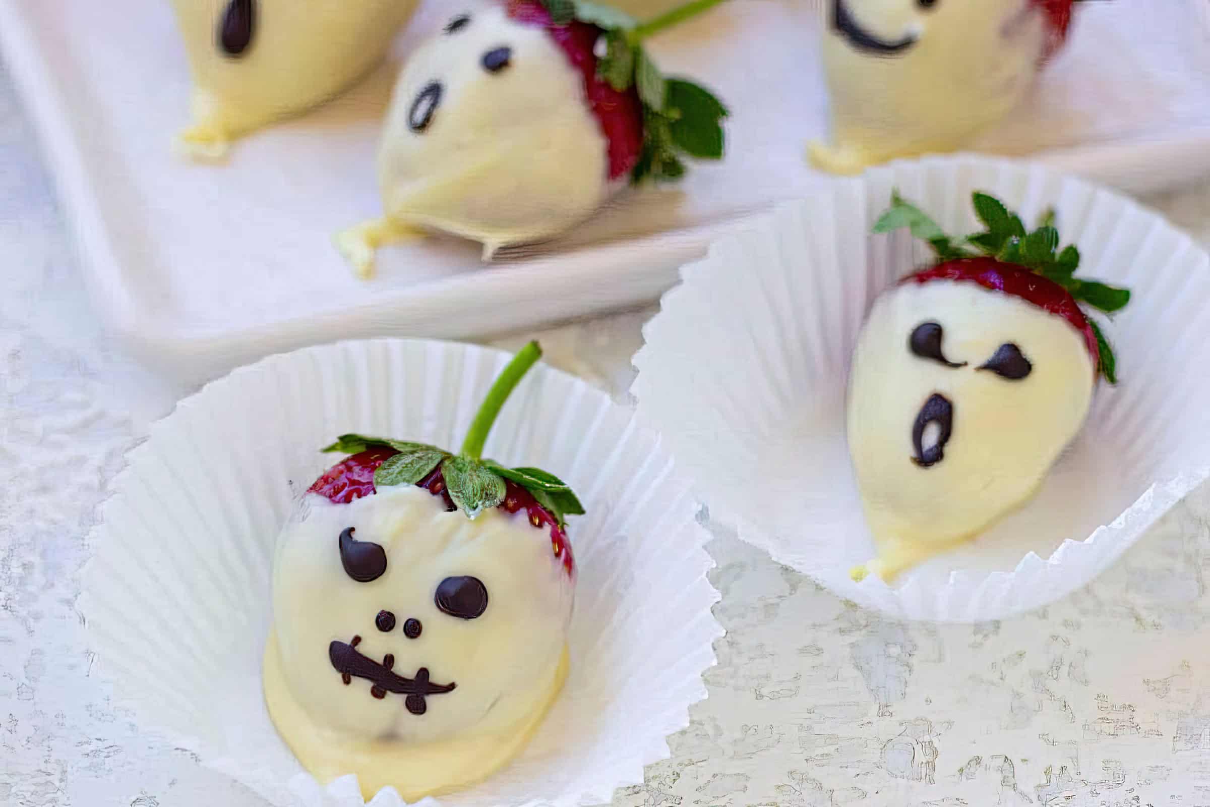 Strawberries coated in white chocolate are decorated with spooky faces using dark chocolate. They are placed in white paper cups on a white surface.