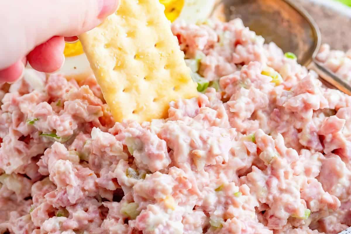 A hand holding a cracker dipped into a bowl of creamy pink ham salad with diced ingredients. A spoon is visible in the background.