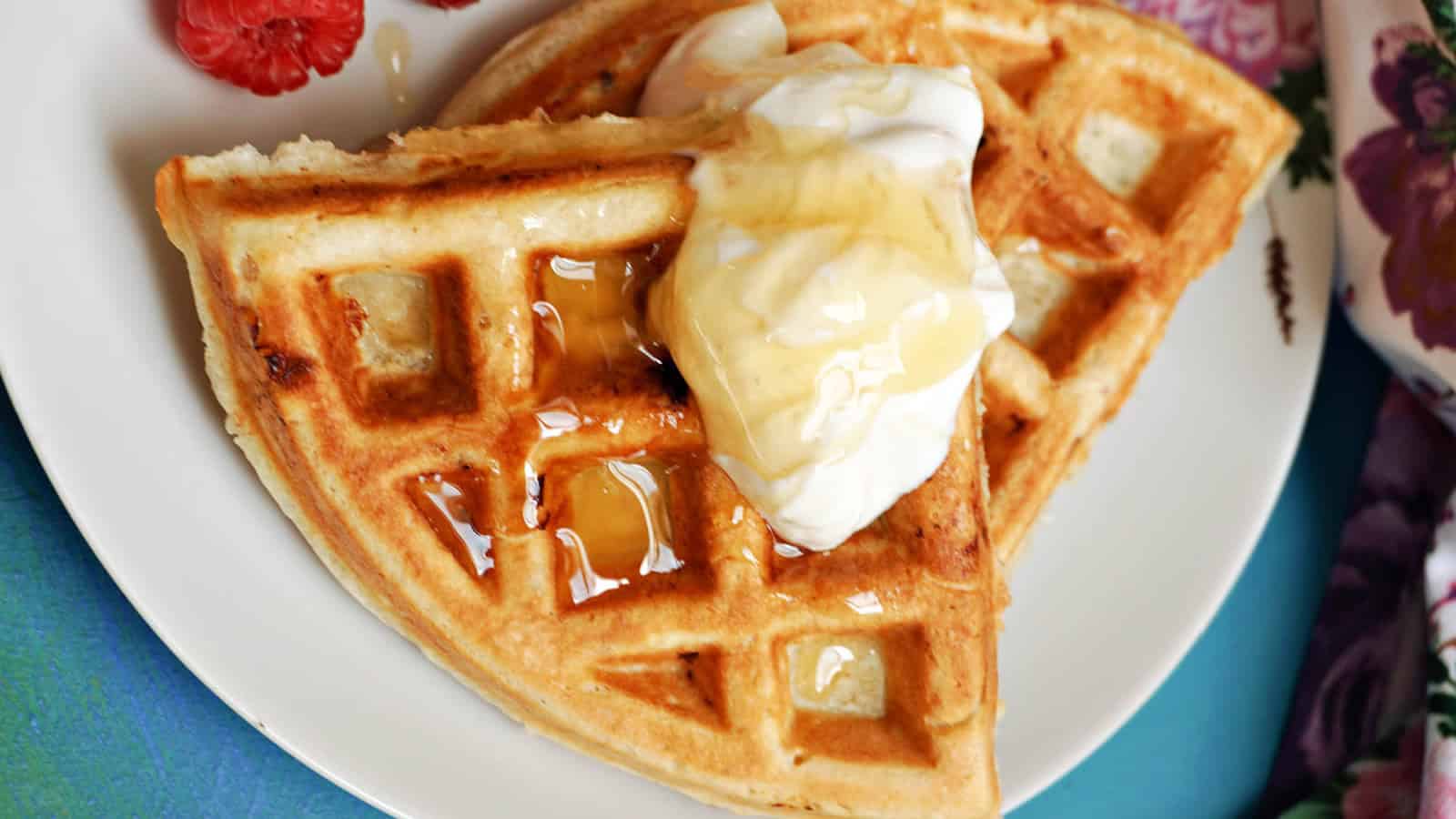 Two golden waffle halves topped with whipped cream and drizzled with syrup on a white plate. A raspberry and part of a floral napkin are visible at the edge.