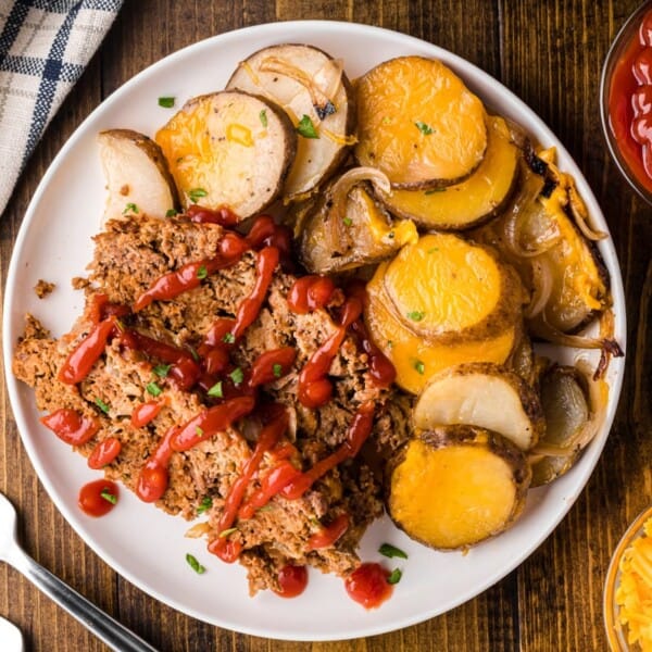 Slices of meatloaf with ketchup, cheesy scalloped potatoes with onions, and shredded cheese on a plate; utensils and napkin nearby.