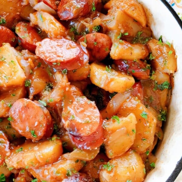 Sliced sausage, potatoes, onions, and herbs arranged in a white pot.
