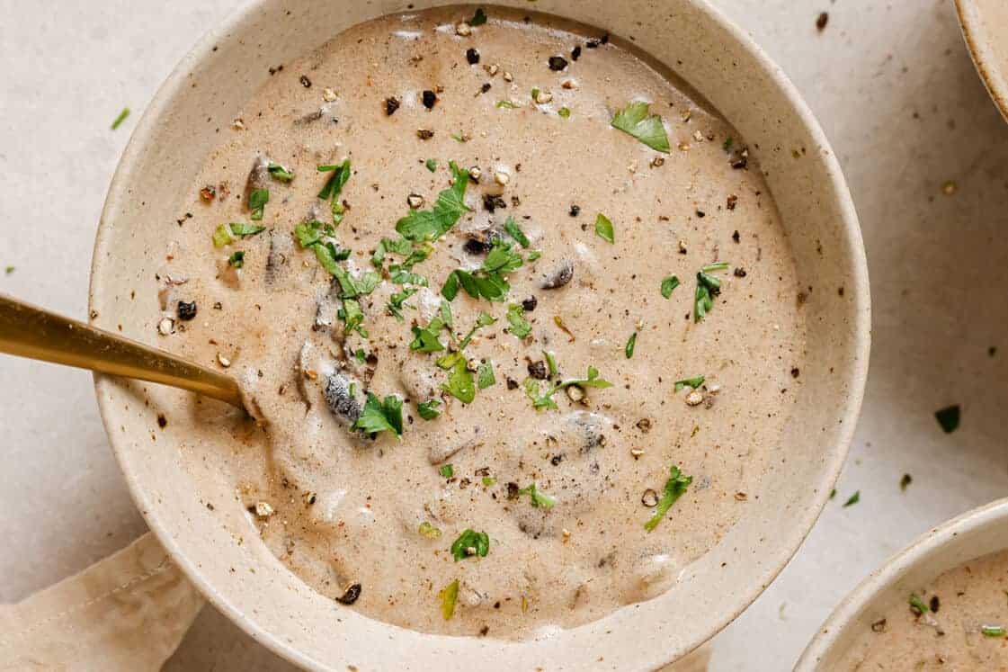 A creamy mushroom soup garnished with chopped parsley and black pepper in a beige bowl. A gold spoon rests inside the bowl. The image is taken from above, showcasing the smooth texture of the soup.