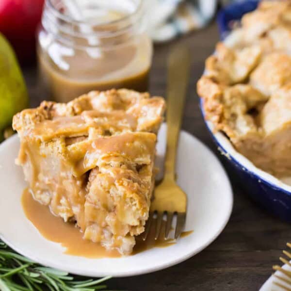 A slice of apple pie with caramel sauce on a white plate with a fork, next to a jar of caramel, an apple, pear, and rosemary.