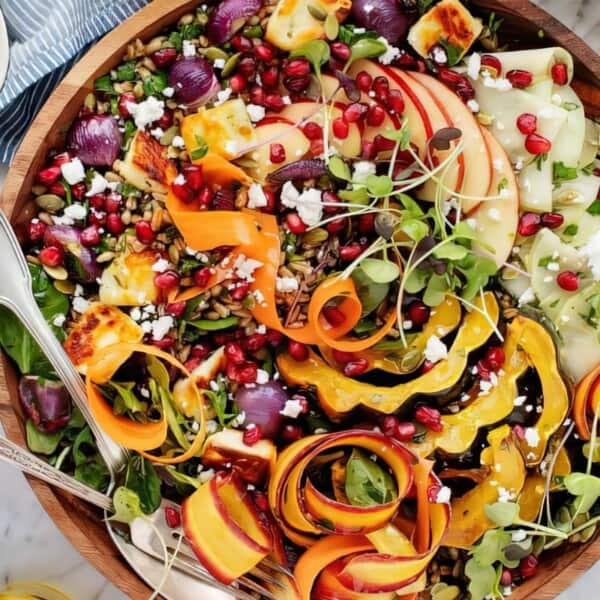 A wooden bowl holds salad with greens, roasted vegetables, sliced apples, pomegranate seeds, feta, microgreens, and utensils.