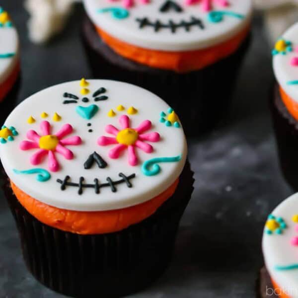 Cupcakes with white fondant, decorated as colorful sugar skulls featuring floral and geometric patterns, on a dark surface.