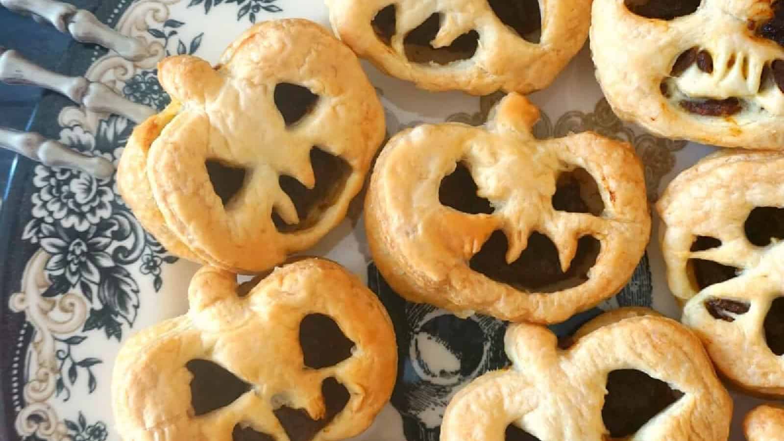 A plate of pumpkin-shaped pastries with smiling faces, resembling jack-o'-lanterns, are arranged on a decorative plate. The pastries have a golden-brown crust and dark filling visible through the cut-out faces.