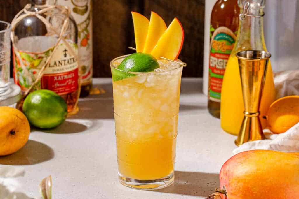 A refreshing tropical cocktail served in a tall glass with ice. Garnished with mango slices and a lime wedge. Surrounded by bottles, a jigger, a whole mango, and vibrant citrus fruits on a countertop.
