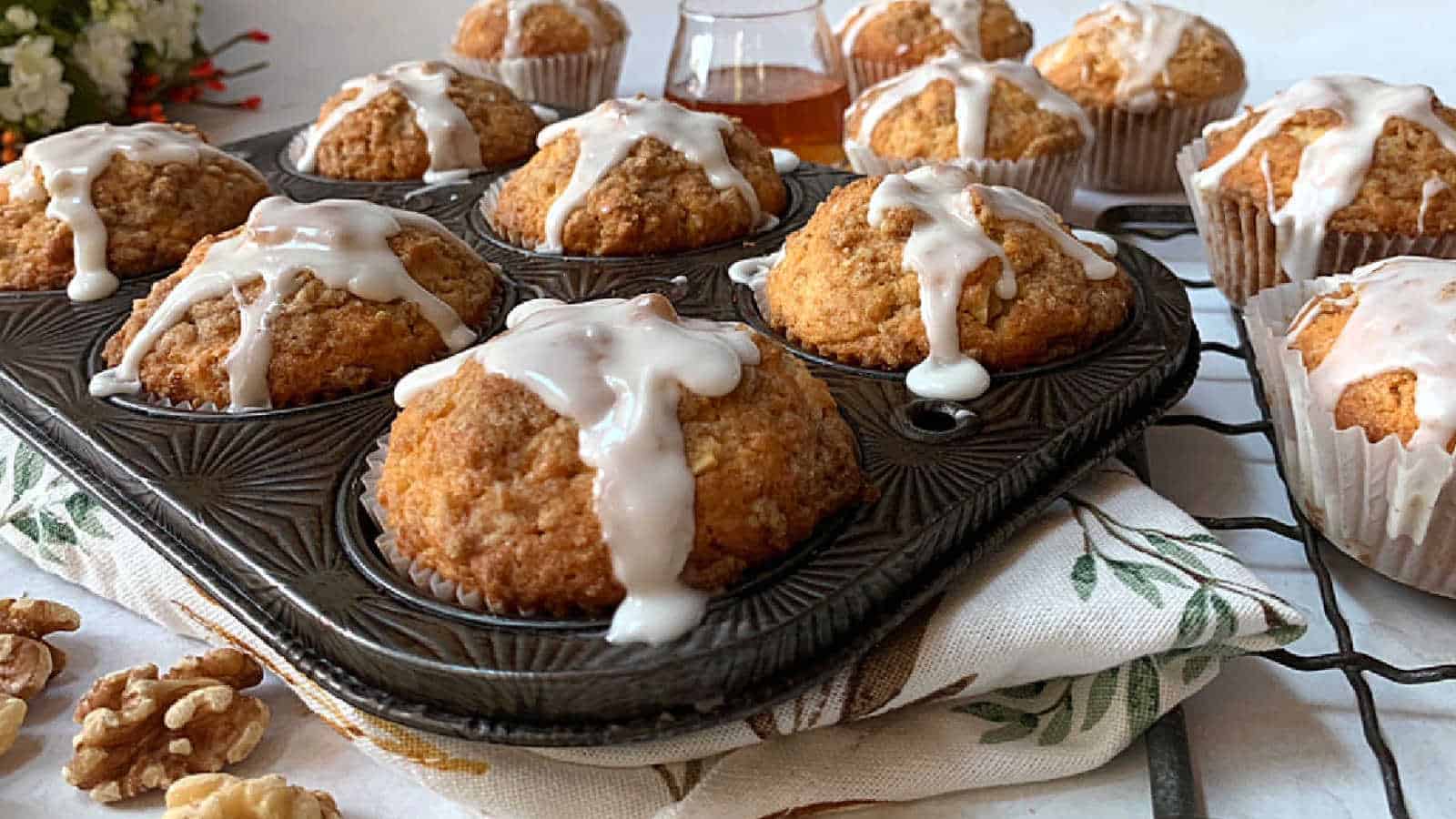 Freshly baked muffins drizzled with icing rest in a dark muffin tray. Some muffins are removed, placed nearby on a patterned cloth. A small glass container with amber liquid sits in the background, and a few walnuts are scattered around.
