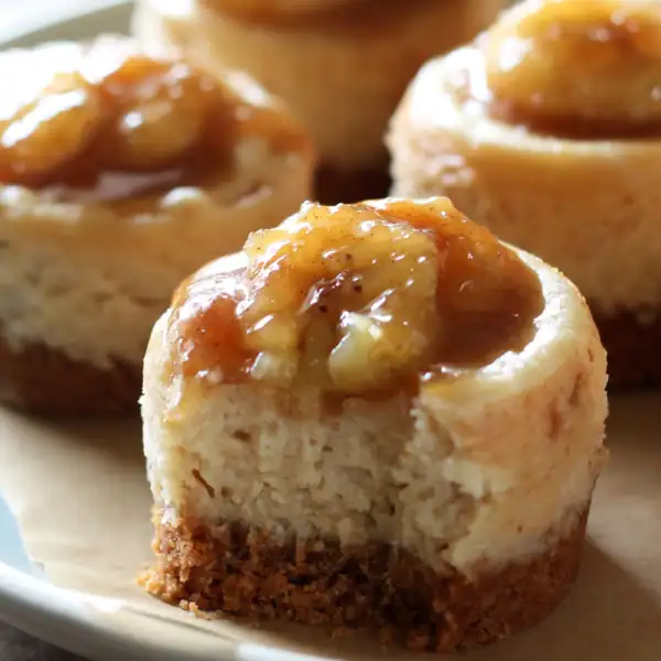 A close-up of mini cheesecakes with crumb crusts, each topped with caramelized banana slices, arranged on a plate.