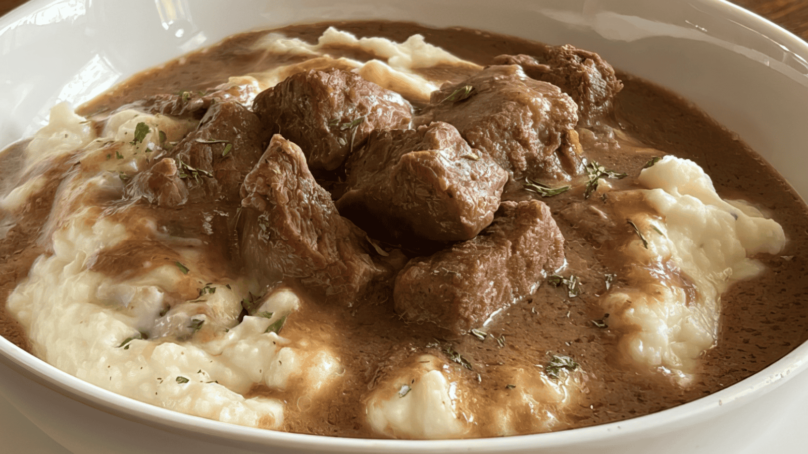 A bowl of creamy mashed potatoes topped with chunks of beef in rich brown gravy, garnished with a sprinkle of herbs.
