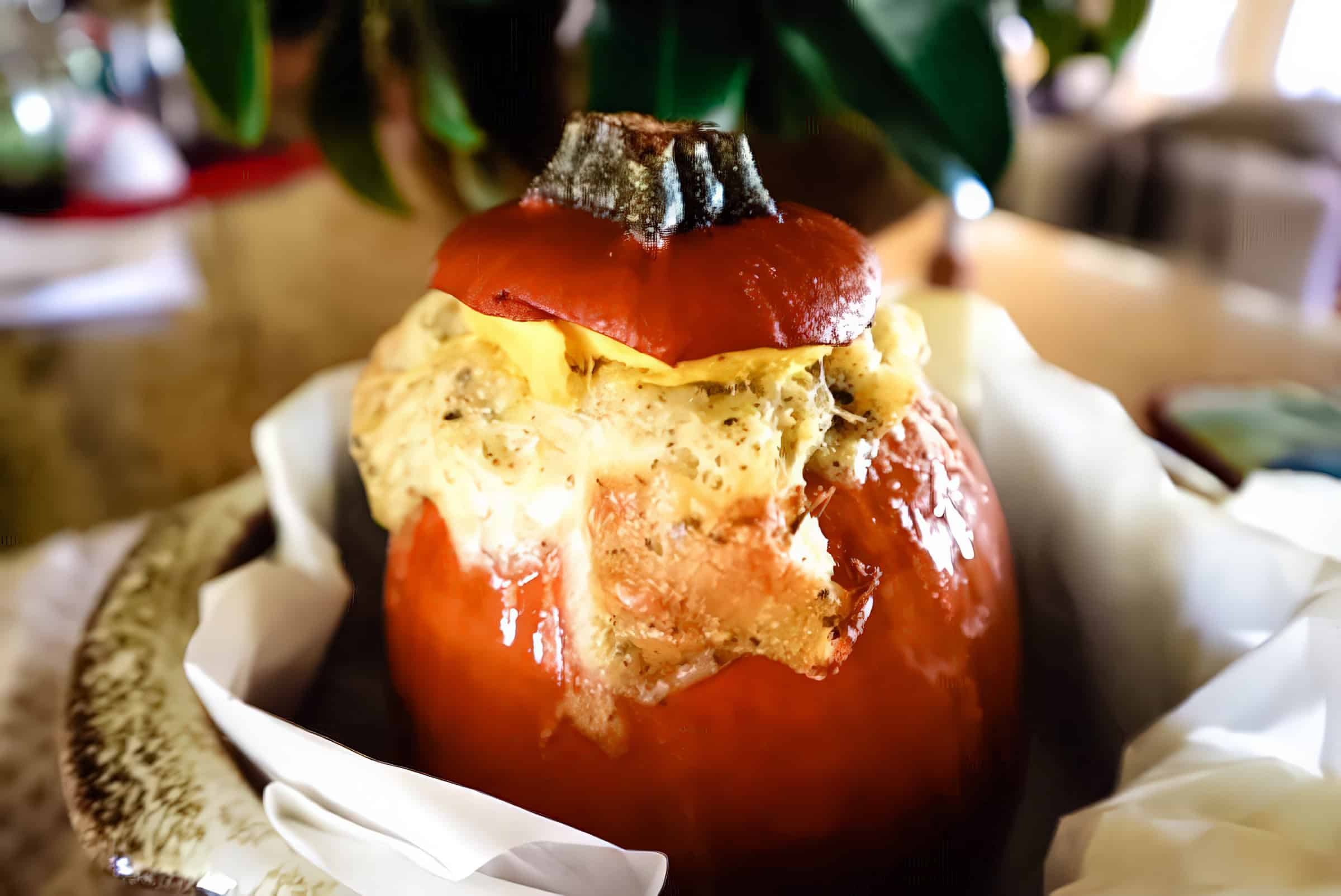 A stuffed pumpkin with a savory filling and melted cheese on top sits on a plate lined with parchment paper. A small pumpkin cap is placed on top. In the background, there's a glimpse of green foliage.