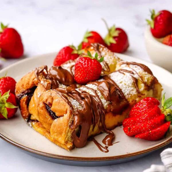 Pastries on a plate with chocolate drizzle and powdered sugar, garnished with strawberries; extra strawberries in a nearby bowl.