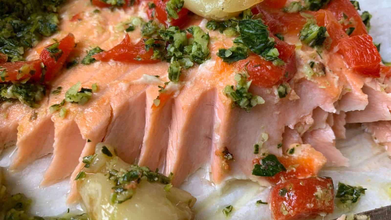 A baked salmon fillet topped with herbs and chopped red peppers, served with roasted garlic cloves.
