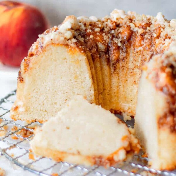 The image features a Bundt cake with a slice removed, topped with crumbly streusel. There's a whole peach placed on a plate in the background. The cake rests on a wire cooling rack.
