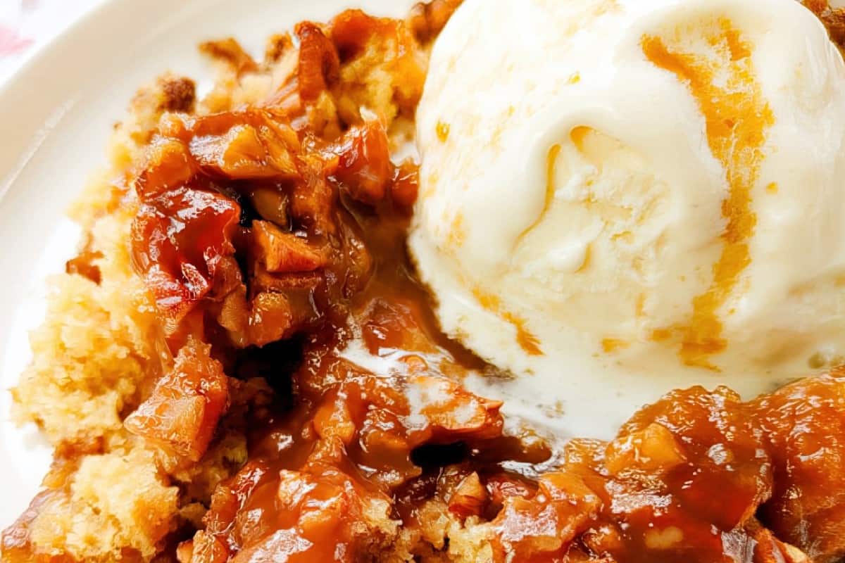 Close-up of a dessert featuring a scoop of vanilla ice cream next to caramelized apple pieces and a crumbly cake. The ice cream is melting slightly, blending with the warm apple topping on a white plate.