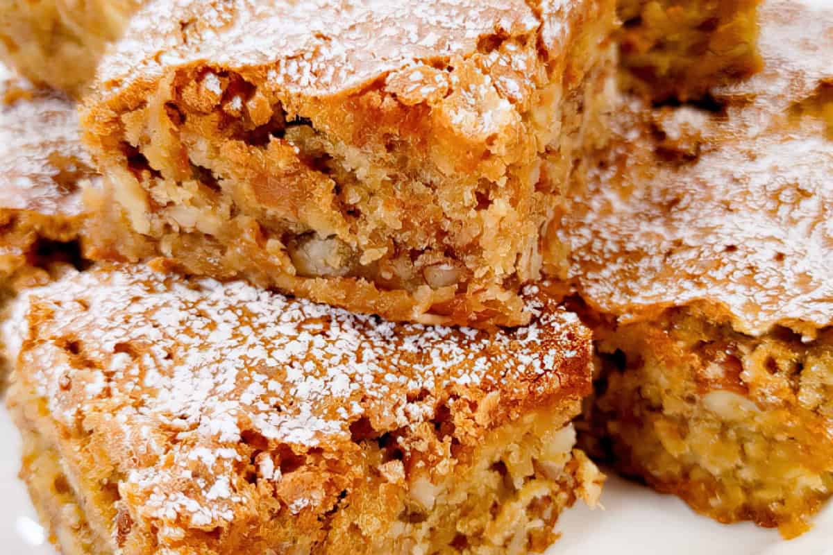 Close-up of several slices of moist apple cake dusted with powdered sugar. The cake is rich in color with a slightly crispy top and visible chunks of apple and nuts, giving it a hearty, homemade appearance.