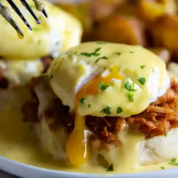 Two eggs Benedict with pulled pork, hollandaise, and parsley beside roasted potatoes; a fork pierces one egg.