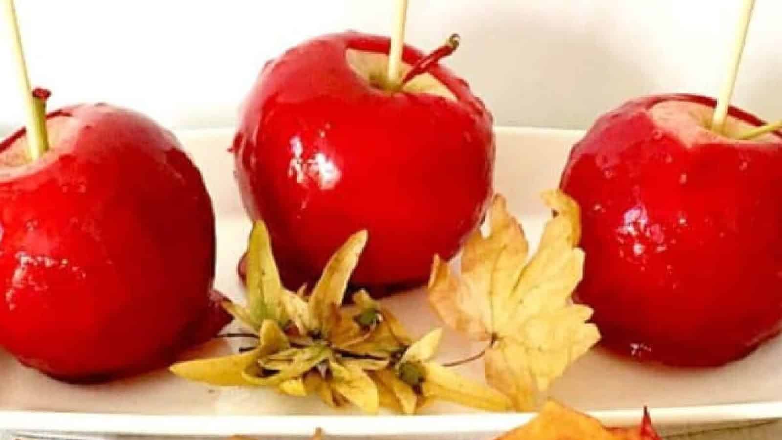 Three bright red candy apples with wooden sticks are displayed on a white plate. Autumn leaves are scattered around them, adding a seasonal touch. The glossy coating on the apples gives them a shiny and appetizing appearance.