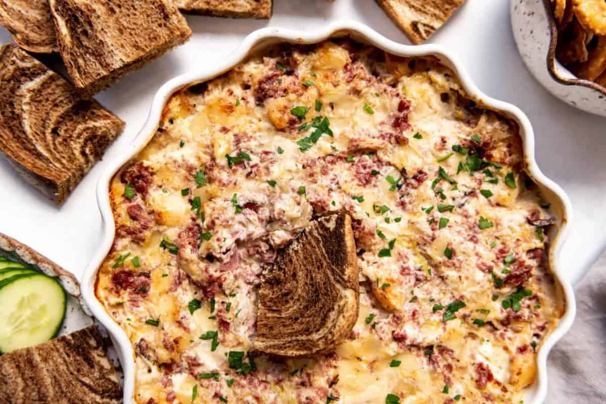 A creamy, baked Reuben dip topped with chopped parsley in a round dish. It is surrounded by slices of marbled rye bread and a few slices of cucumber on the side.