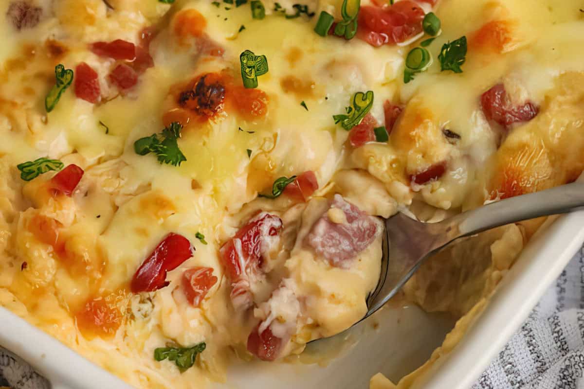 A close-up of a creamy casserole dish with melted cheese, topped with chopped tomatoes and herbs. A spoon is scooping out a portion, revealing layers of vegetables and possibly meat beneath the cheese.