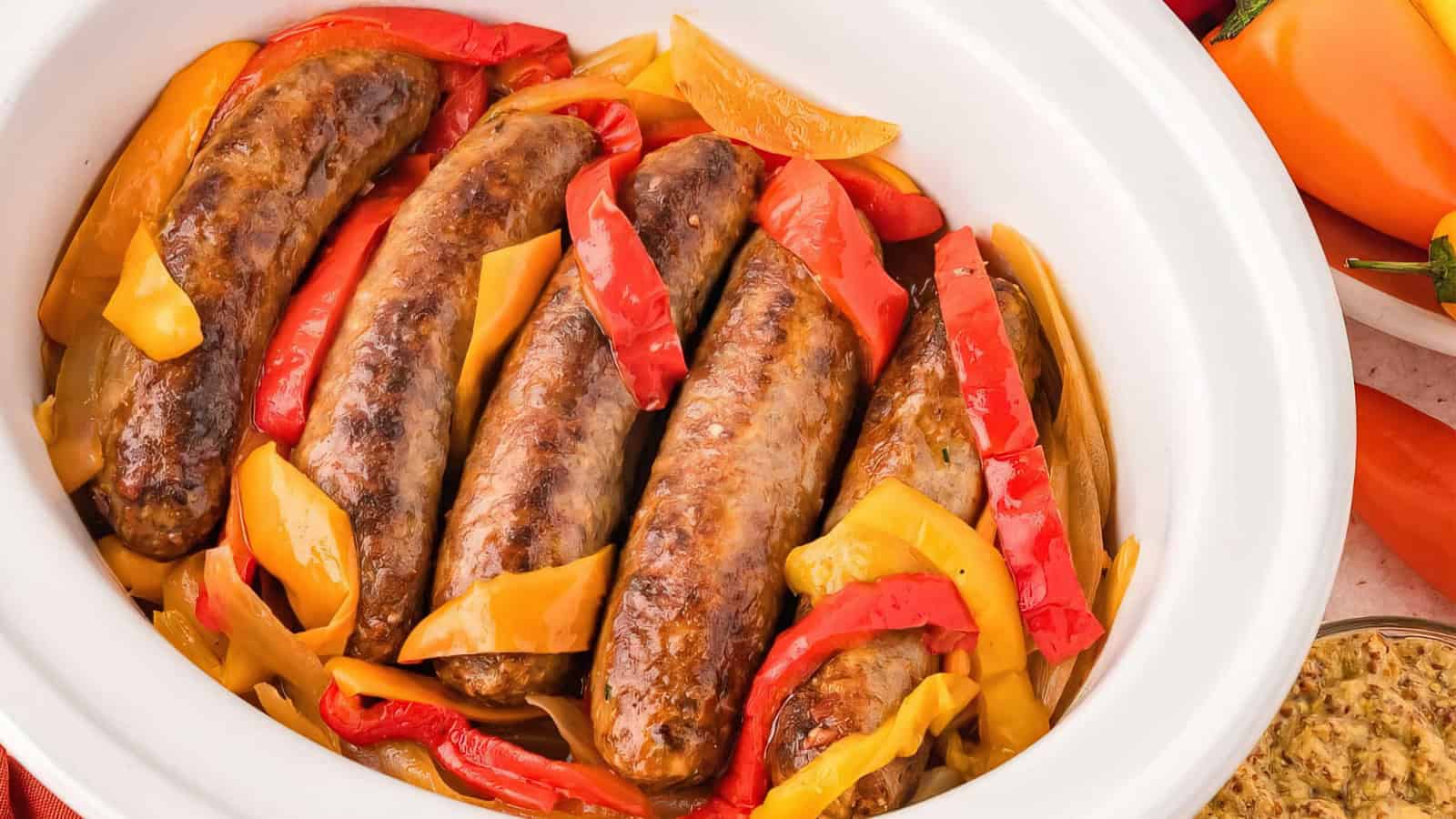 A white slow cooker filled with cooked sausages nestled among sliced red and yellow bell peppers. The colorful dish is vibrant and inviting, surrounded by fresh bell peppers in the background.