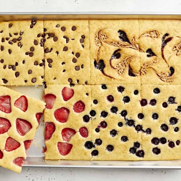 A baking sheet with pancake squares topped with chocolate chips, strawberry slices, blueberries, and swirled spreads; one square lifted.