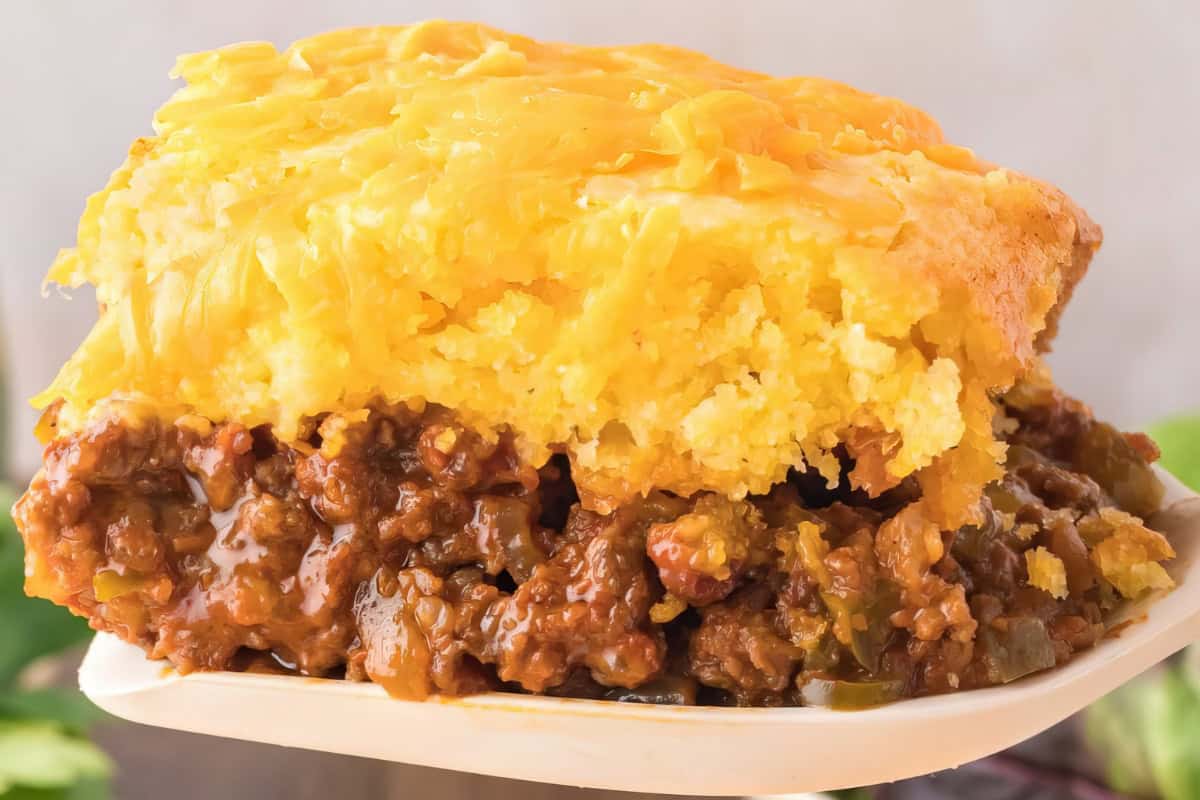 A close-up of a slice of savory pie with a layer of golden cornbread topping and a filling of ground beef mixed with vegetables and sauce. The pie slice rests on a light-colored plate.