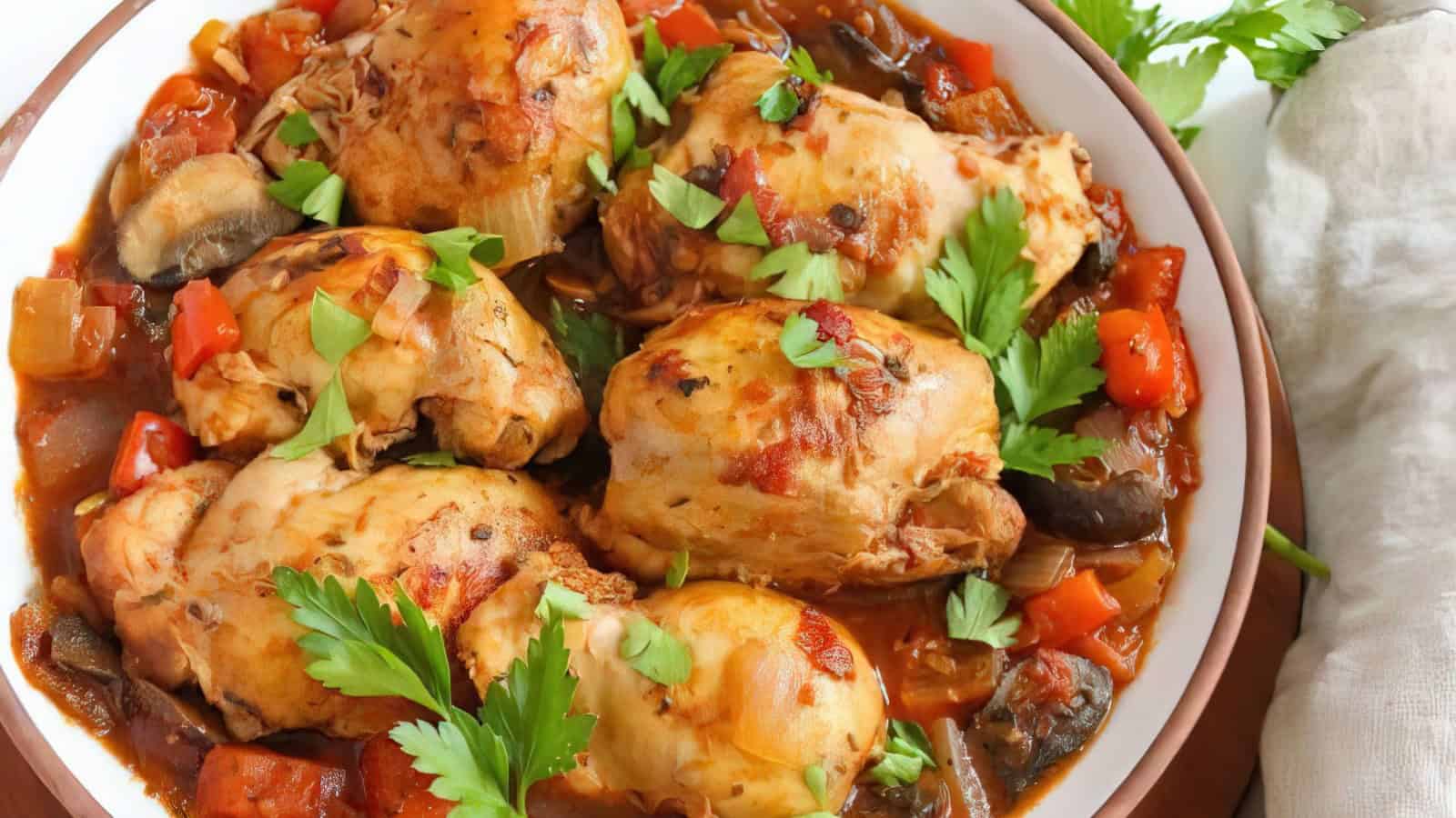 A bowl of chicken cacciatore with cooked chicken pieces, tomatoes, peppers, and mushrooms, garnished with fresh parsley leaves.
