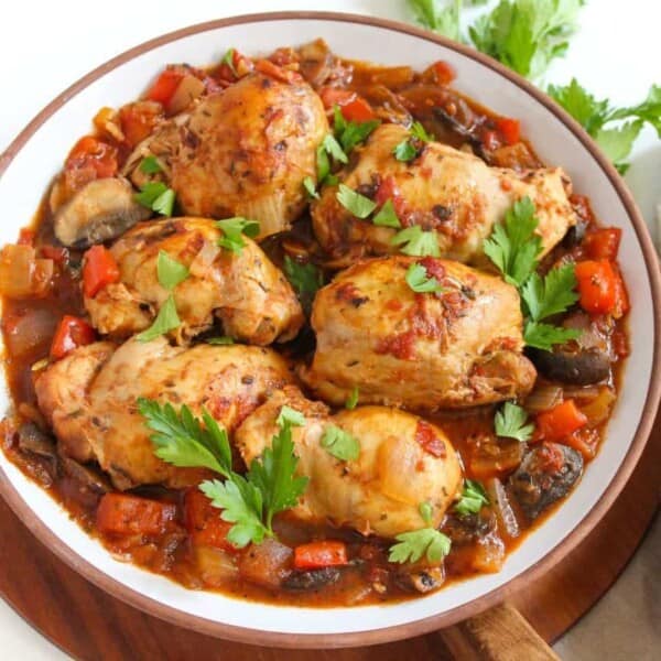 Cooked chicken pieces topped with parsley next to a stew containing tomatoes, mushrooms, onions, and peppers on a plate.