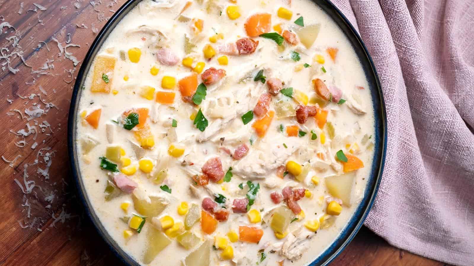 A bowl of soup containing chicken chunks, potatoes, corn, carrots, and herbs is on a wooden surface next to a pink cloth.