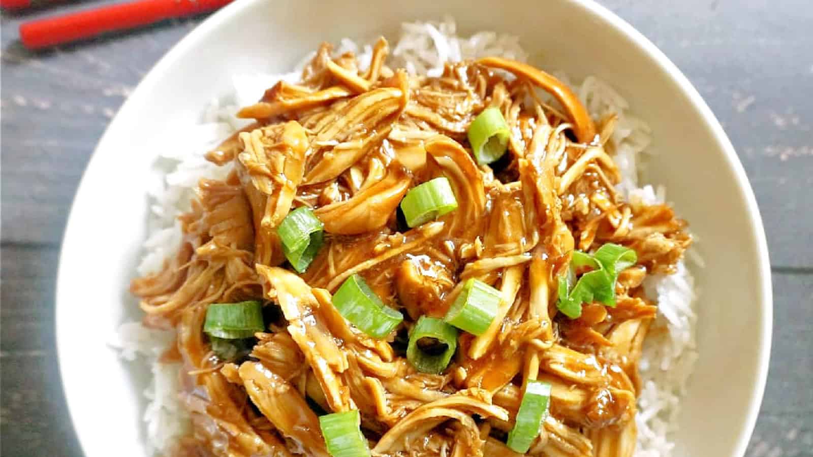 A bowl of shredded chicken covered in a brown sauce, garnished with chopped green onions, served on a bed of white rice. The dish is presented on a wooden table with a pair of red chopsticks visible in the background.