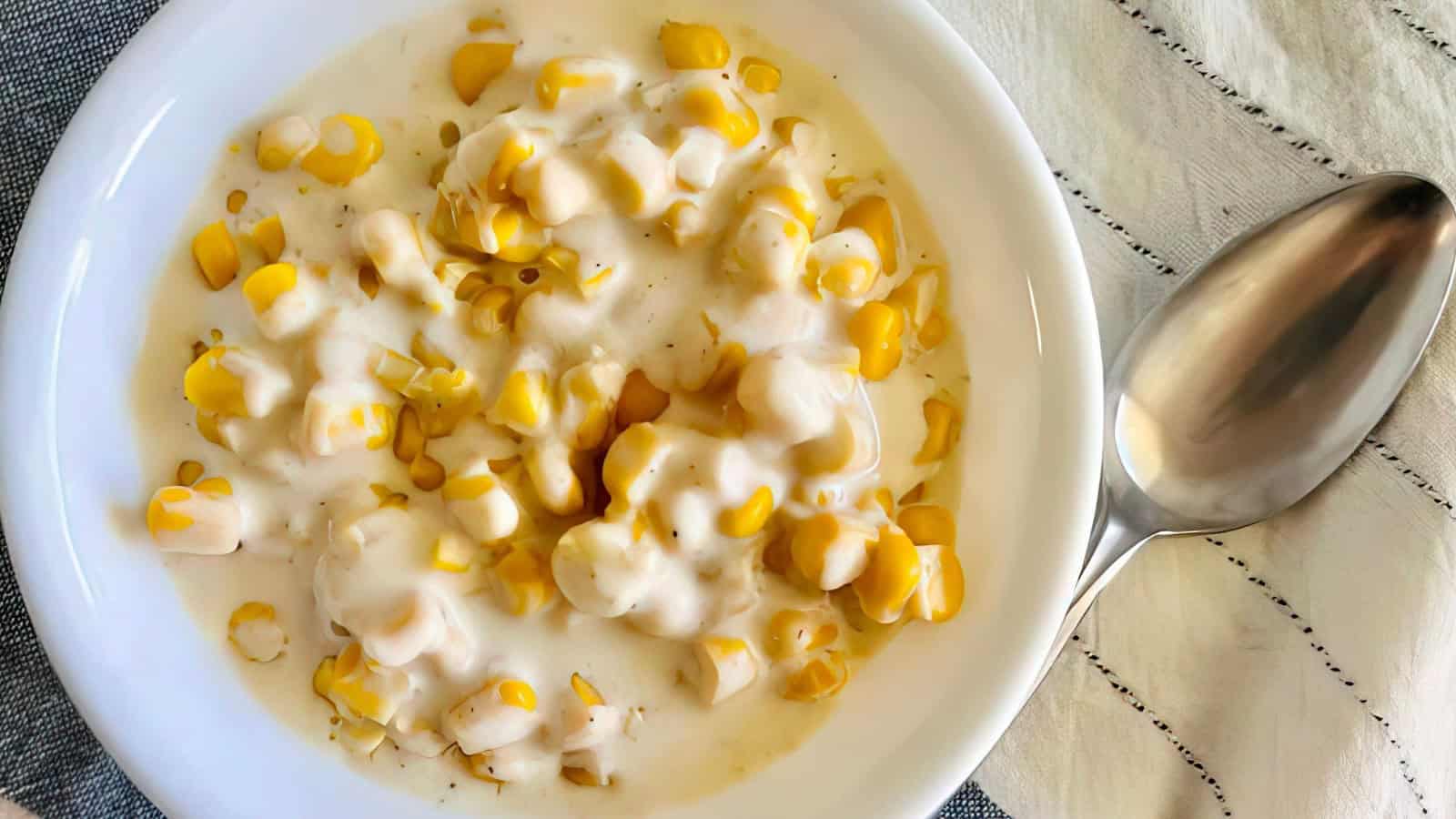 A white bowl filled with creamy corn sits on a cloth napkin. To the right of the bowl, there is a stainless steel spoon. The corn is covered in a thick, white sauce.