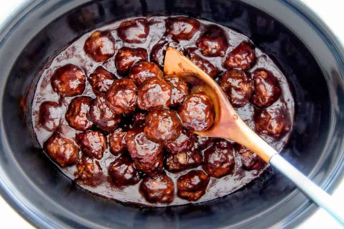 A slow cooker filled with meatballs in a rich, dark sauce. A wooden spoon rests to the side, partially submerged in the sauce.