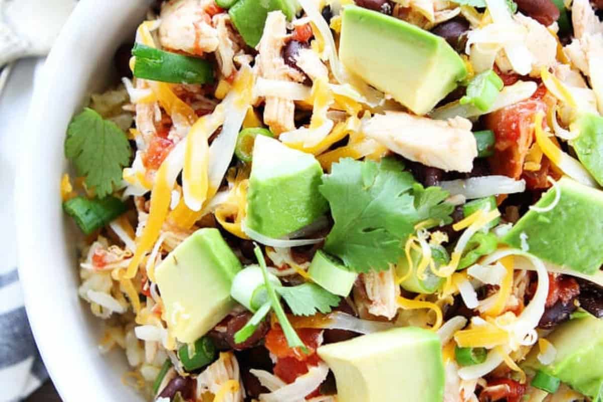 A colorful salad featuring diced avocado, shredded cheese, black beans, chicken pieces, and chopped cilantro, all mixed with red diced tomatoes and sliced green onions, served in a white bowl.