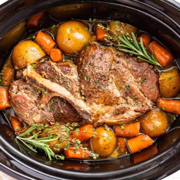 A cooked pot roast with potatoes, carrots, and sprigs of rosemary and thyme in a black slow cooker.