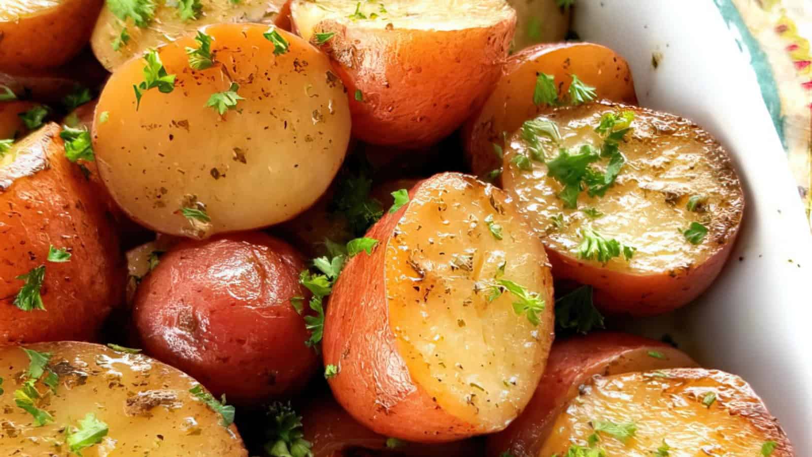 Close-up of halved roasted red potatoes garnished with fresh parsley and seasoned with herbs, displayed in a white dish. The potatoes have a golden-brown, crispy exterior.