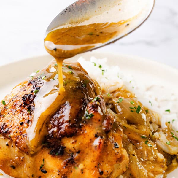 A spoon pours brown sauce over cooked chicken on a plate with white rice, garnished with herbs.
