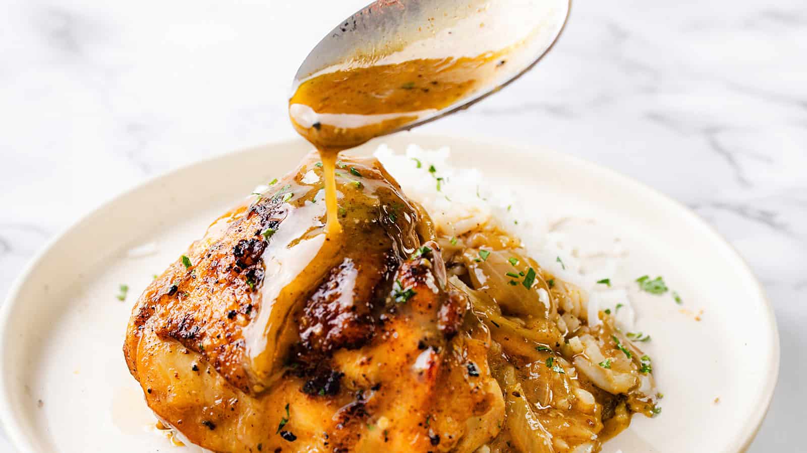 A spoon drizzles savory sauce over a piece of grilled chicken served with a side of white rice and garnished with herbs on a white plate. The dish is placed on a marble surface.