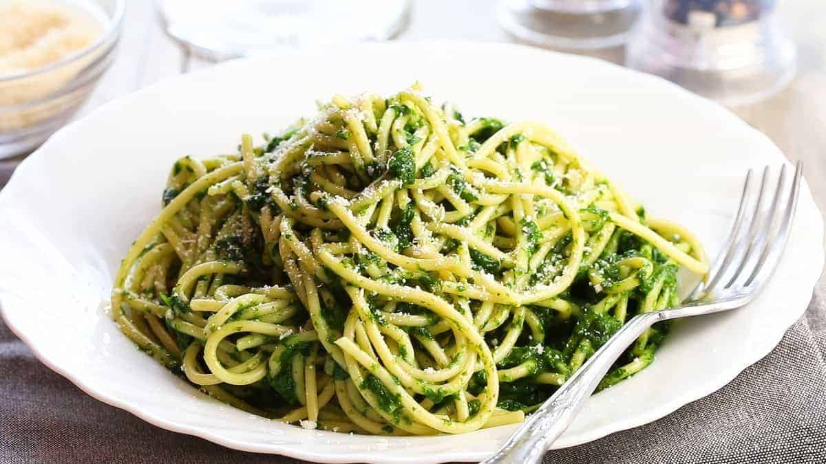 A white plate with spaghetti tossed in a vibrant green pesto sauce, sprinkled with grated cheese. A fork rests on the edge of the plate. The background includes blurred glassware and a gray napkin.