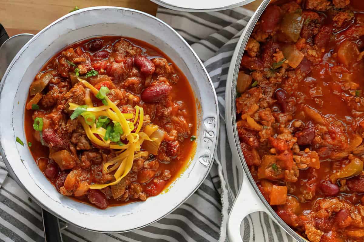A bowl of chili with minced meat, kidney beans, tomatoes, and diced peppers, topped with shredded cheddar cheese and fresh herbs. The bowl is placed on a striped cloth next to a pot filled with more chili.