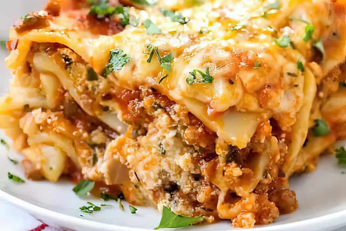 Close-up of a cheesy lasagna slice on a white plate, featuring layers of pasta, meat, tomato sauce, and melted cheese. Fresh herbs are sprinkled on top for garnish.
